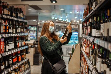 Young cheerful woman in medical face mask shopping during pandemic standing in wine store