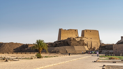 A paved sidewalk leads to the ancient Egyptian temple of Horus in Edfu. High stone walls with...