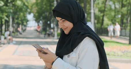 Happy muslim woman using smartphone in city park on sunny day outdoors. Smiling muslim woman wearing hijab headscarf typing texting message on phone in city street. Communication in social networks