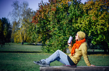 Happy woman enjoying coffee on park bench on green foliage background. Autumn concept. Leisure time in sunny fall day. Copy space.