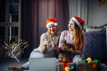 Fototapeta premium Two happy women in red Santa Claus hats using laptop and communicating remotely with friends and family. New Year decorations and cute kitten in home interior.