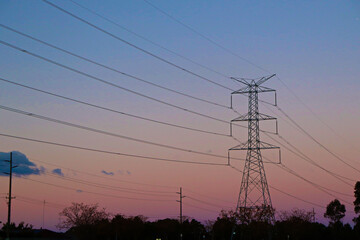 Large overland power lines spanning across suburban wetlands area toward mountains in the distance, with the sun setting in the background.