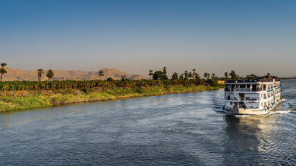 A cruise ship is sailing on the blue river. On the shore there is green vegetation, palm trees....