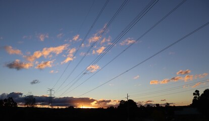 Large overland power lines spanning across suburban wetlands area toward mountains in the distance, with the sun setting in the background. - Powered by Adobe