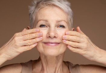 Portrait of mature beautiful woman massaging her face isolated on brown background