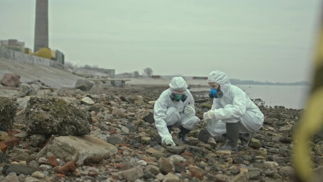 Slowmo shot of female scientists in protective coveralls exploring polluted seashore area and taking soil sample putting it into test tube