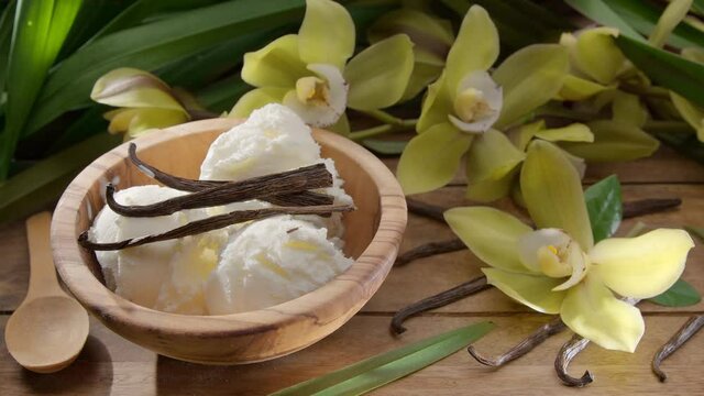 Vanilla ice cream balls in a bowl with vanilla orchid flowers and vanilla sticks lying nearby on a vintage wooden table with green leaves in the back. Slow panoramic camera movement.