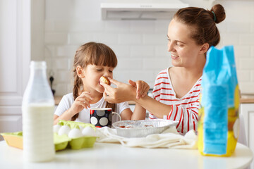 Obraz na płótnie Canvas Indoor shot of optimistic attractive mother wearing striped shirt posing with her female kid and eating homemade cakes after baking, mommy giving to bite pie to her child.