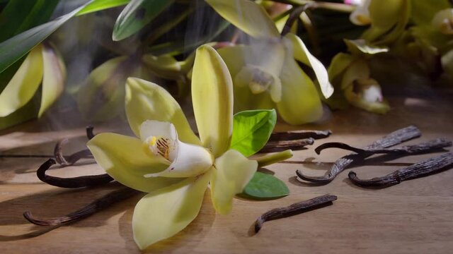 Vanilla orchid flower with vanilla sticks on a vintage wooden table with green leaves in the background. Vanilla gives off a "scent" in the form of small smoke clouds. Slow panoramic camera movement.