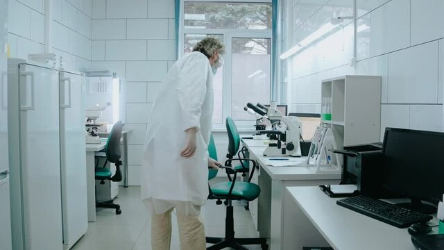 Laboratory assistant sits on a chair and works with a microscope in white lab. An adult male is working in laboratory with test tubes wearing mask. High quality 4k footage