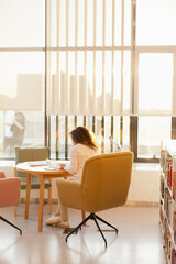 young beautiful woman sitting at the table on chair using mobile phone at home near big windows.  Woman using mobile at the university library or office.