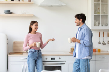 Fototapeta premium Satisfied smiling millennial woman and guy with cups of hot drink, have breakfast, talking on kitchen interior