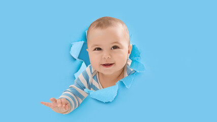 Smiling cute adorable european little kid climbs through hole on blue paper, copy space, panorama