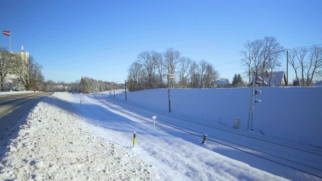 Latvian flag by roads in Sigulda in snowy winter. Snow-covered railway tracks next to an asphalt road. The national flag of Latvia over the building of the bobsleigh track. Gauja National Park in