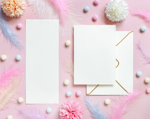 Blank cards and envelope near pastel flowers, pom-poms and feathers near ring in a gift box on pink