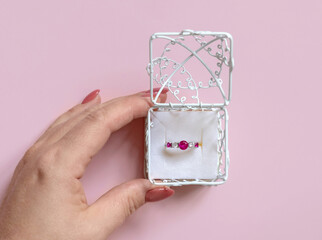 Womans hand holding a ornate gift box with a gemstone engatement ring on pink