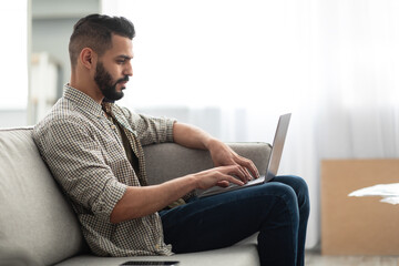 Online work, communication. Young Arab guy making video call, browsing internet on laptop, sitting on couch at home