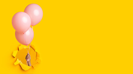 Hand of millennial african american lady holding three pink balloons through hole in yellow paper, panorama - Powered by Adobe