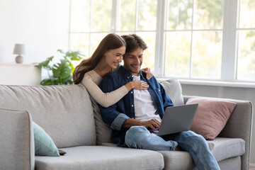 Naklejka na ściany i meble Happy cheerful millennial european female hugs husband sit on sofa, man works on laptop in living room