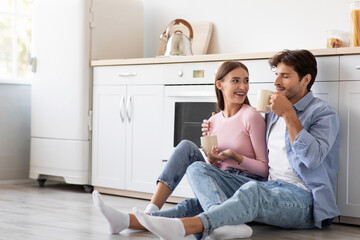 Satisfied glad young european lady and guy with cups of favorite drink sits on floor, talk in kitchen