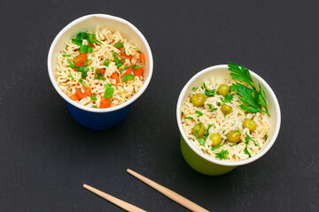 Prepared Instant Noodles with Green Pea, Carrot and Greens in Blue and Green Cups with Chopsticks on Black Background. Quick Lunch or Unhealthy Fast Food