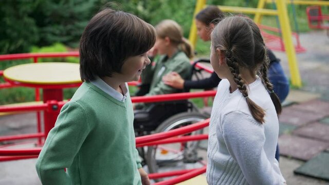 Caucasian boy and girl mocking laughing at blurred disabled girl on wheelchair at background. Rude children sneering on playground in park outdoors. Discourtesy and childhood