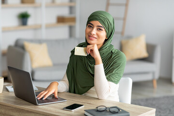 Online shopping. Young Arab woman sitting at her desk with laptop, using credit card to buy goods...