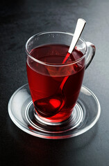 Red fruit tea in tea glass with glass coaster, on black concrete surface.