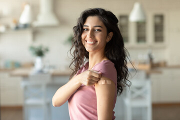 Cheery young brunette female showing arm with band aid after coronairus vaccine injection at home