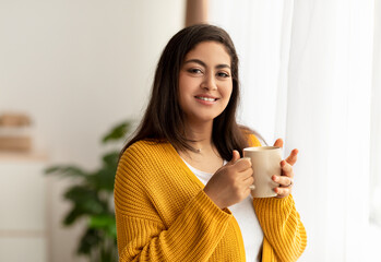 Portrait of happy arab lady standing near window with cup of hot beverage, smiling at camera, free space