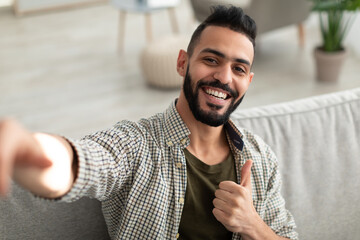 Happy young Arab guy taking selfie and showing thumb up gesture, smiling at mobile phone camera,...
