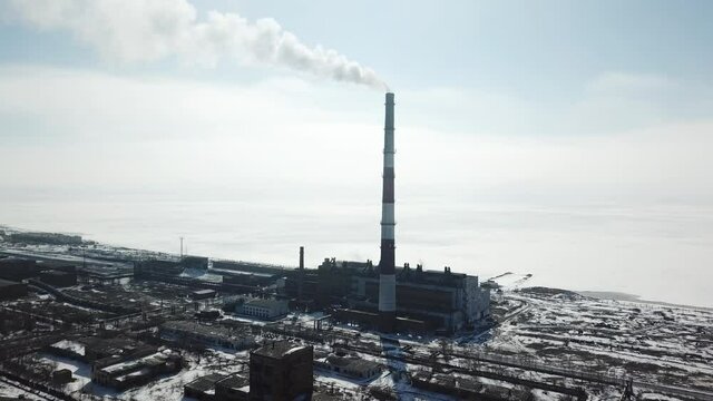Moving aerial shot around snowed heat and power plant and its smoking chimney when weather is sunny. Iced big water behind plant on the right of frame, some buildings on the left. Blue sky