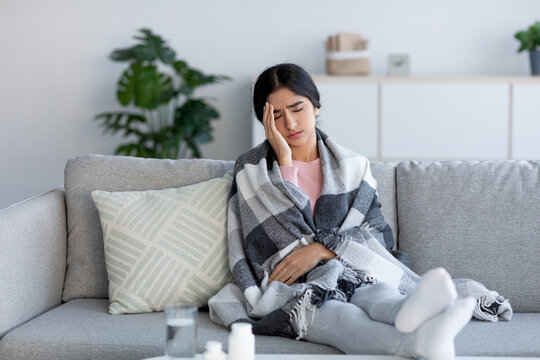 Tired sad upset young hindu lady wrapped in plaid, suffering from headache and flu, sits on sofa