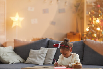 Portrait of cute African-American girl writing letter to Santa while sitting at table in cozy living room with Christmas tree in background, copy space