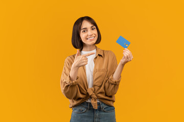 Positive young lady pointing at credit card recommending banking service, posing over yellow background and smiling