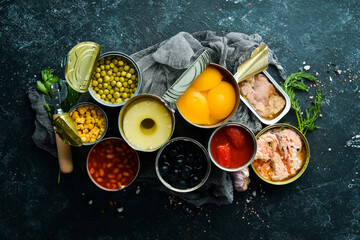 Canned vegetables, beans, fish and fruits in tin cans on black stone background. Food stocks.