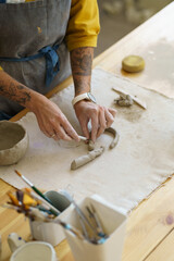 Cropped photo of craftswoman potter standing by table in studio trimming and modeling mug handles, preparing clay material for pottery, female hands making ceramics during masterclass or workshop