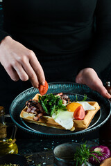 Sandwich with egg, tomatoes and bacon on a plate in the hands of a chef On a black background. Belgian waffles.