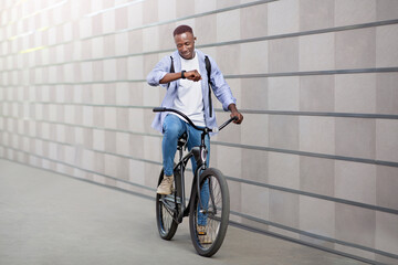 Happy African American guy with earphones listening to music and checking time on smartwatch during bike ride outside