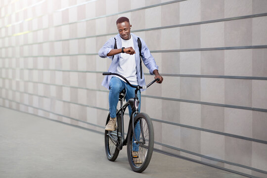 Happy African American guy with earphones listening to music and checking time on smartwatch during bike ride outside