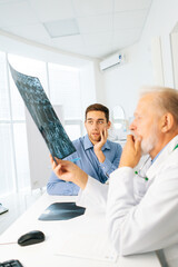 Obraz premium Vertical shot of mature adult male physician consulting scared young man patient giving bad news explaining results of MRI image. Unhappy frustrated young man listening to bad news, looking at camera.