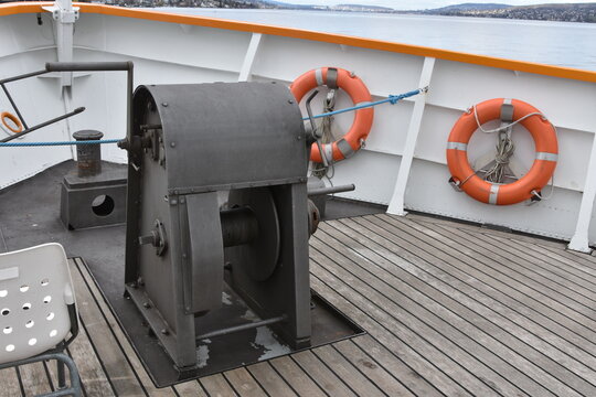 Anchor gear fixed on wooden boarder floor in bow of a tourist cruiser ship painted white. On main weather bottom deck is also installed double bollard and white plastic chair and two orange life buoys