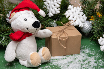 A Christmas gift in a craft box and a Teddy bear in a red Santa hat and a scarf on a table with snow and Christmas decorations