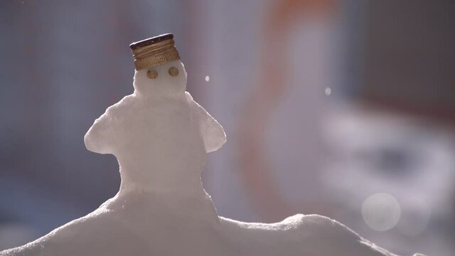 Close up of little snowman with pea eyes and bottle metal cap on head standing on snowdrift when sunny and falling drops. Snowman is on the left of frame, background is out of focus