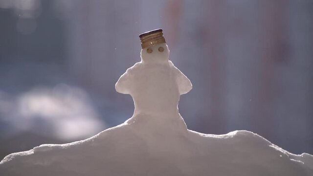 Little snowman with pea eyes and bottle metal cap on head standing on snowdrift when sunny and falling drops. Snowman is in the center of the frame, city building is on out of focus background