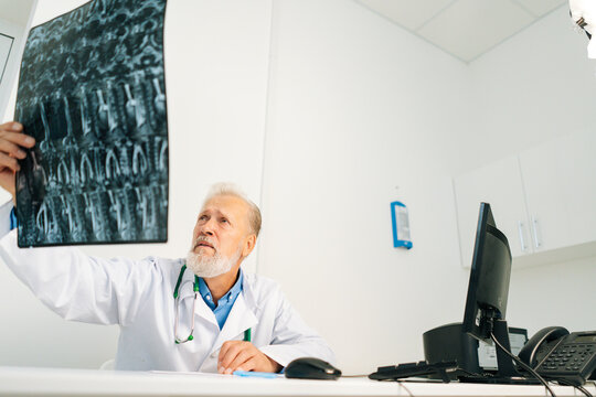 Low-angle view of serious senior adult male doctor examining MRI spine image of patient sitting at desk with computer in medical office room, thinking about diagnosis, expressing concern of illness.