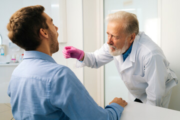 Close-up view back to mature adult male doctor testing sick young man patient for possible coronavirus infection using throat swab in medical office. Man doing PCR test during corona virus epidemic.