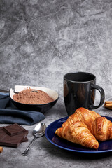Three fresh croissants on a blue plate next to a bowl with cocoa powder and a cup, some chocolate chips, cinnamon sticks and a napkin