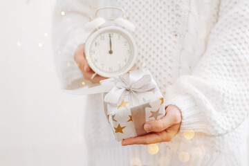 A girl in a white sweater holds a gift and an alarm clock. Holiday concept New Year. Selective focus, light background,