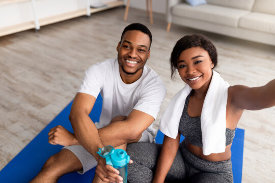 Millennial black couple in sportswear sitting on yoga mats, taking selfie together on smartphone after domestic training